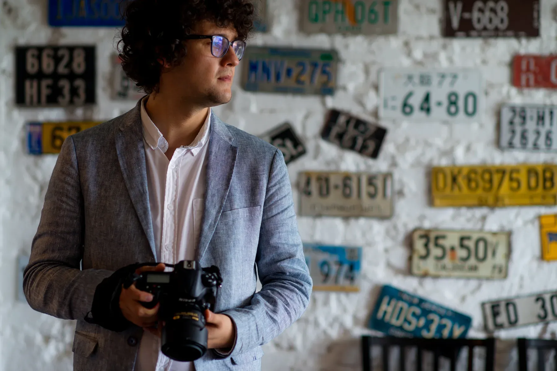 Marius Dragan in a grey blazer holding his camera, looking to the side, in front of a white wall covered in old licence plates